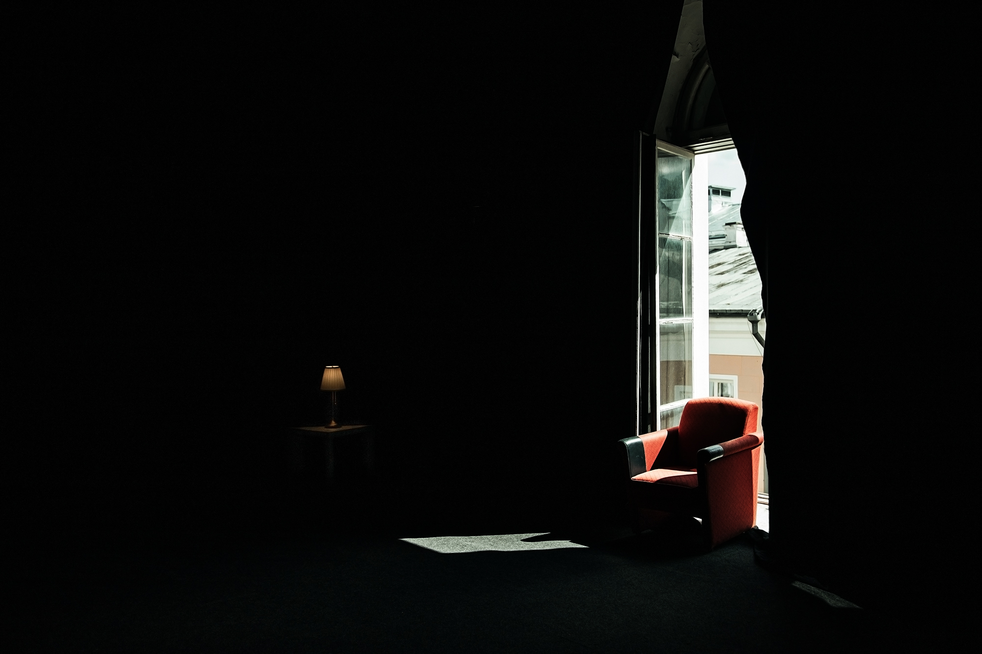 A dark room with a red armchair beside an open window, illuminated by daylight.