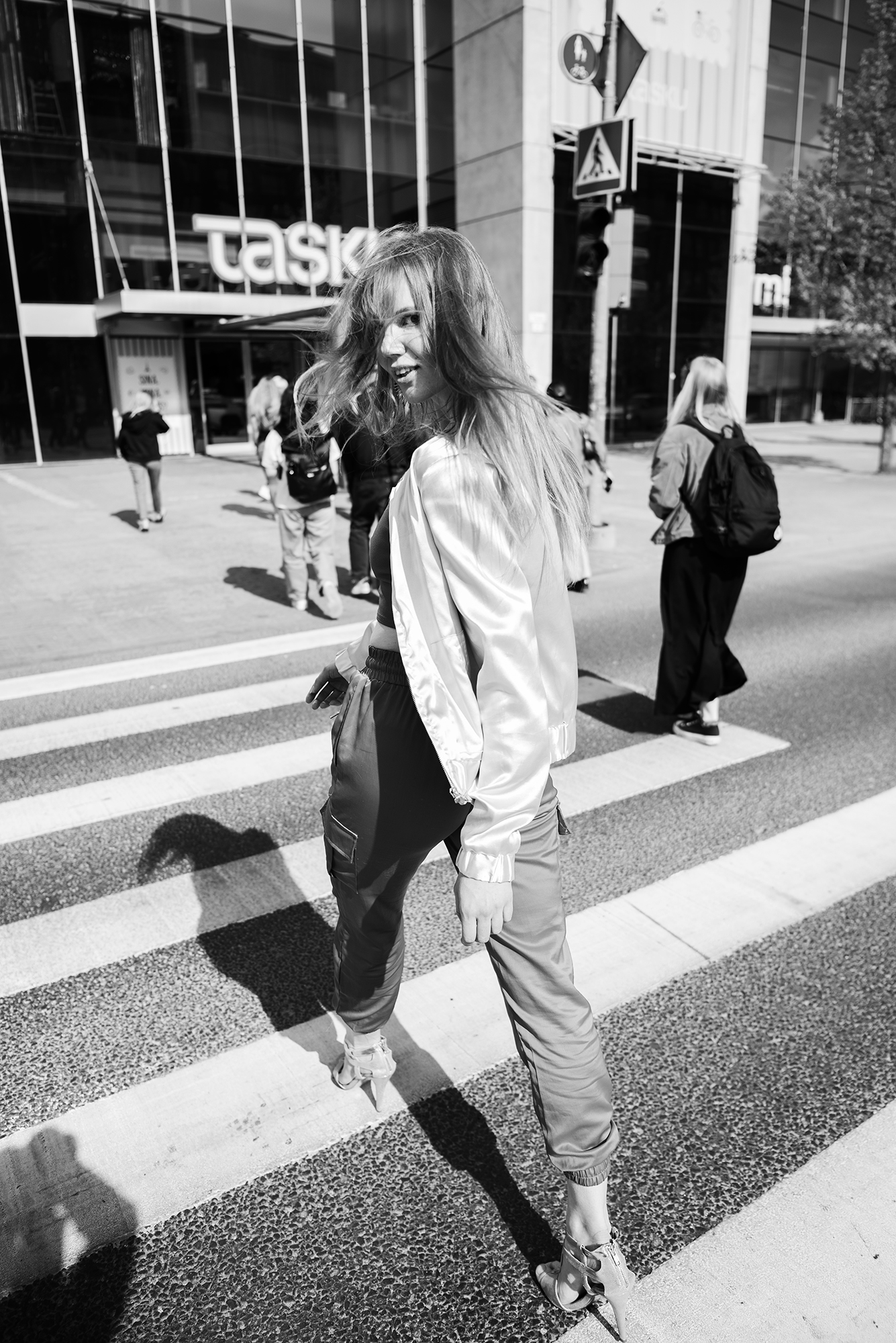 Black and white photograph of Helen Müürsep crossing a city street in a satin bomber jacket, hair caught in the wind